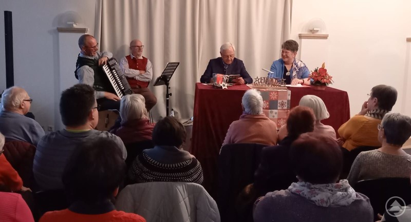 Akteure des Advents-Nachmittags von links: Alois Göppel, Martin Ertl, Walter Körner und Traudl Wagner Vier Akteure sitzen vorne an einem Tisch und musizieren bzw. lesen Adventsgeschichten vor einem aufmerksamen Publikum in einem Lesungssaal. Zwei Männer spielen Akkordeon und Gitarre, zwei weitere Personen lesen Geschichten. Der Raum ist gut besucht, die Stimmung ist heiter und gemeinschaftlich.