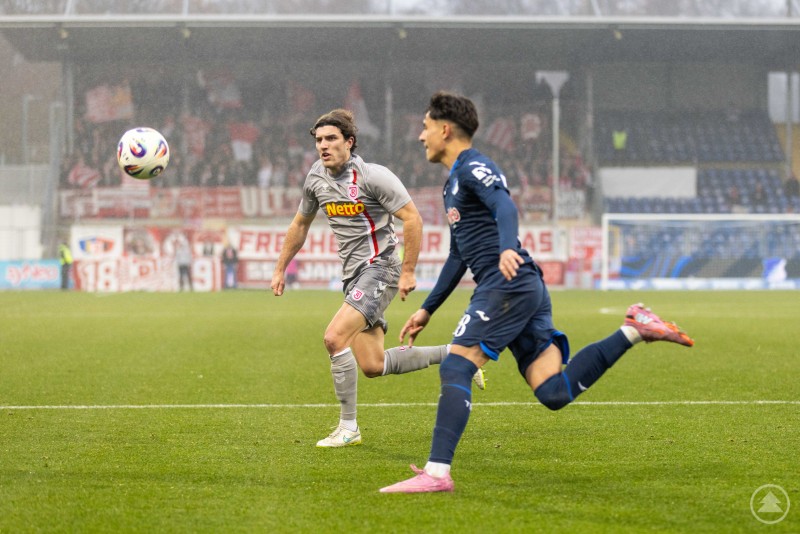 Laufduell zwischen einem Regensburger und einem Hoffenheimer Spieler. Ein Jahn-Spieler sprintet dem Ball hinterher, ein Hoffenheimer Spieler läuft neben ihm.