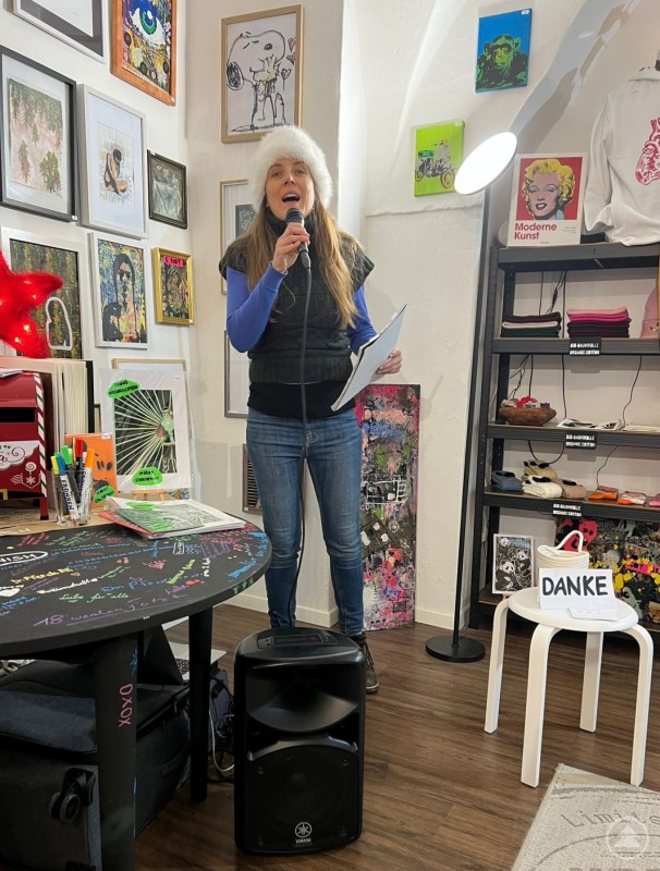 Die Sopranistin Christina Harslem Eine Frau steht in einem kleinen, kunstvoll gestalteten Raum und singt in ein Handmikrofon. Sie trägt eine helle Mütze, eine dunkle Weste und Jeans und hält ein Blatt Papier in der Hand. Um sie herum hängen zahlreiche bunte Kunstwerke und Illustrationen an den Wänden; im Hintergrund sind Regale mit weiteren Bildern und Gegenständen zu sehen. Die Szene wirkt intim und erinnert an ein kleines Konzert oder eine Lesung in einer Galerie.