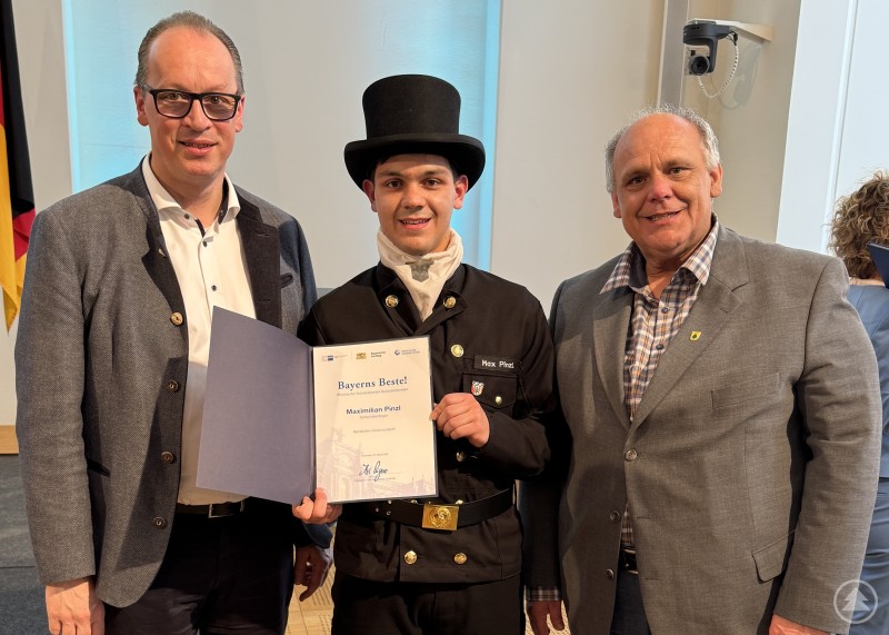 Bei der Ehrung Bayerns Beste 2026 im Maximilianeum gratulierte MdL Martin Behringer dem ausgezeichneten Schornsteinfeger Maximilian Pinzl persönlich zu seinem bundesweiten Erfolg. Drei Männer stehen bei einer Ehrung im Maximilianeum nebeneinander. Die Person in der Mitte trägt die Berufskleidung eines Schornsteinfegers mit Zylinder und hält eine Urkunde in der Hand.
