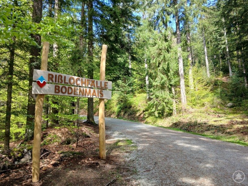 Holzschild mit der Aufschrift „Rißlochfälle Bodenmais“ am Waldrand, Wanderweg im Hintergrund.