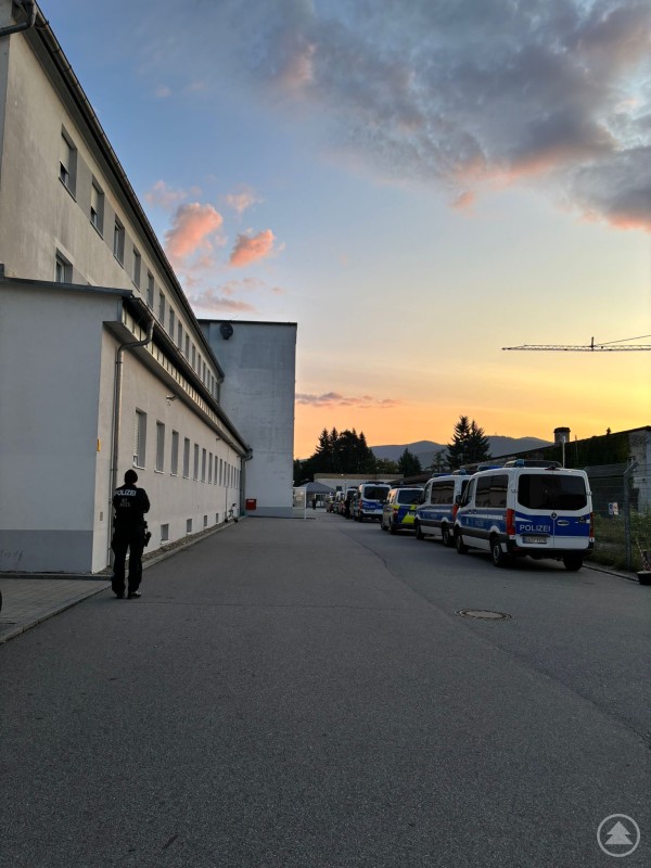 Bereits in den frühen Morgenstunden begann der Polizeieinsatz an der Unterkunft in Deggendorf. Ein Polizist steht neben einer Reihe von Polizeifahrzeugen bei Sonnenaufgang vor einem langgestreckten Gebäude.