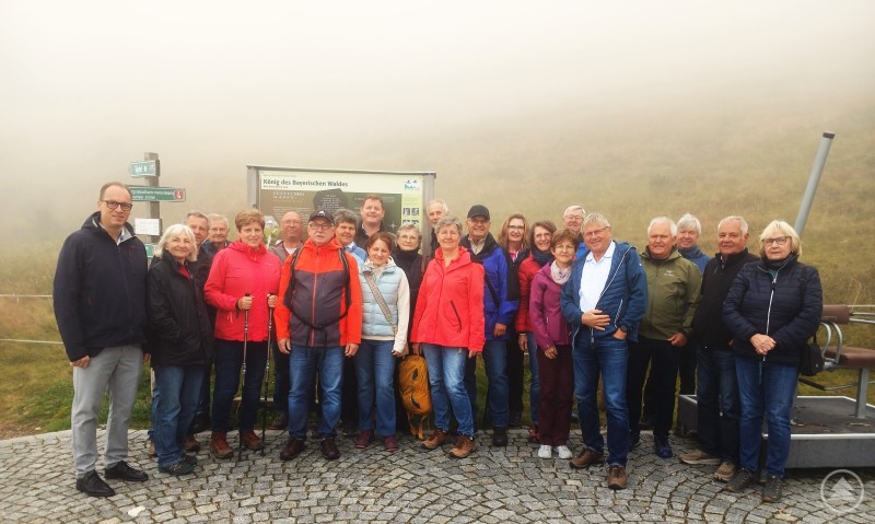 Kolpingsfamilie Regen mit Organisatorin Marianne Wartner (6. v. l.) und MdL Martin Behringer (1. v. l.) bei ihrem Besuch der Radarstation am Großen Arber. Eine Gruppe von Männern und Frauen steht in Wanderkleidung im Nebel auf dem Großen Arber, vor einer Infotafel mit der Aufschrift „König des Bayerischen Waldes“.