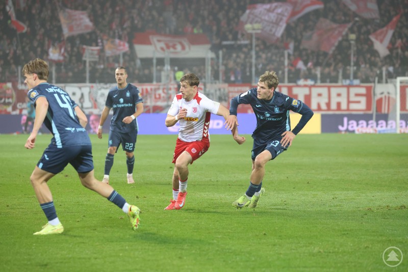 Zweikampfstark und fokussiert – der SSV Jahn Regensburg dominierte über weite Strecken das Duell gegen 1860 München. Ein Spieler des SSV Jahn Regensburg läuft mit dem Ball nach vorne, eng bedrängt von einem Gegenspieler des TSV 1860 München. Im Hintergrund sind weitere Spieler und jubelnde Fans mit rot-weißen Fahnen zu sehen.
