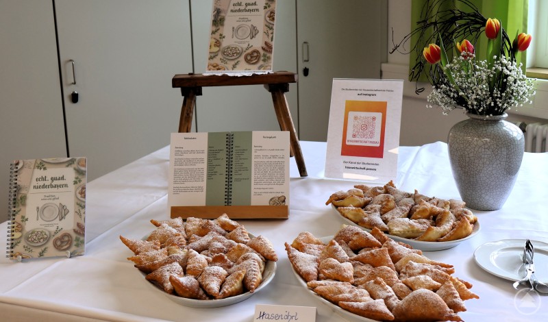 Ein selbst erstelltes kleines Kochbuch soll die alten Rezepte aus dem Landkreis vor dem Vergessen bewahren. Ein Buffet mit traditionellen Gebäckstücken, daneben ein aufgeschlagenes Kochbuch und ein Schild mit QR-Code.