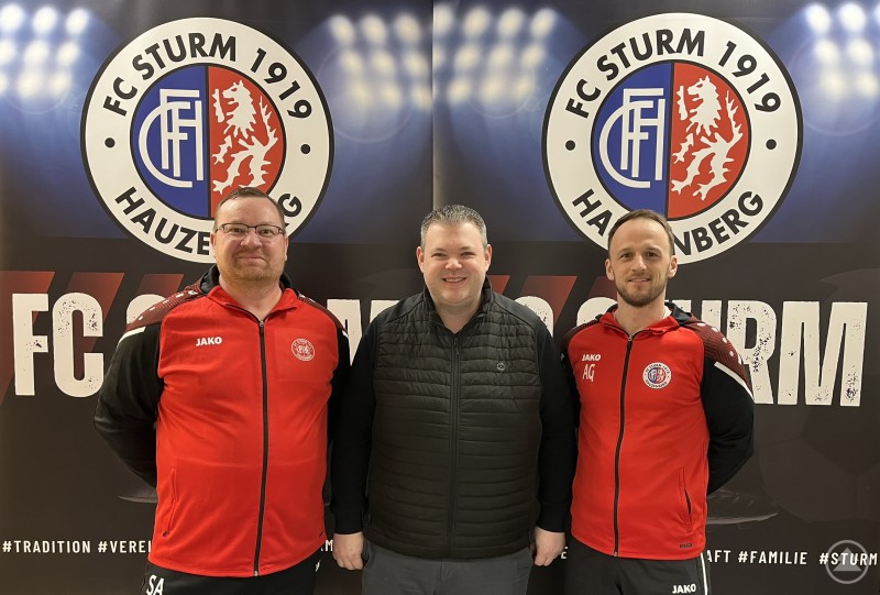 Der FC Sturm Hauzenberg stellt die sportliche Leitung neu auf: Alexander Geiger übernimmt ab der Saison 2026/27 die Rolle des Sportkoordinators, Stefan Anetzberger wird Teammanager der ersten Mannschaft. Drei Männer stehen vor einer Vereinswand des FC Sturm Hauzenberg. In der Mitte posiert ein Mann in dunkler Jacke, links und rechts daneben stehen zwei Männer in roten Vereinsjacken.
