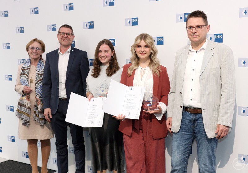 Bei der IHK-Auszeichnung in Passau wurden auch zwei Prüfungsbeste aus dem Landkreis Freyung-Grafenau geehrt. Fünf Personen stehen vor einer IHK-Fotowand. Zwei junge Frauen halten ihre Urkunden in den Händen und werden gemeinsam mit drei weiteren Personen für ihre Leistungen gewürdigt.