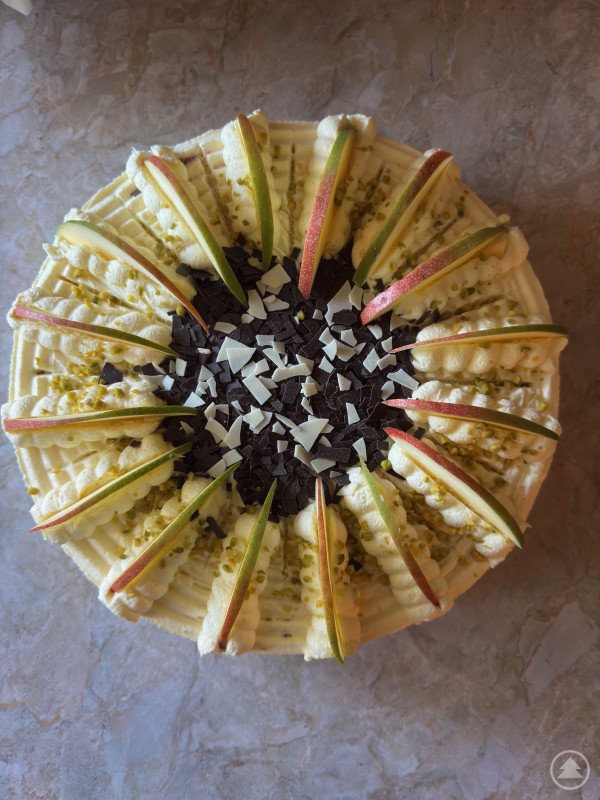 Eine dekorative Torte mit Sahnetupfen, Apfelscheiben, Pistazien, dunklen und weißen Schokoraspeln auf einem hellen Untergrund.