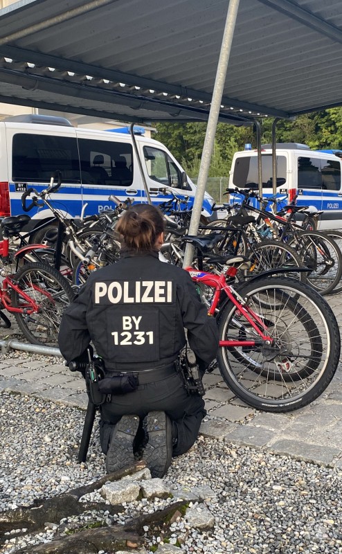 Eine Beamtin kontrolliert im Rahmen des Einsatzes Fahrräder auf dem Gelände der ANKER-Einrichtung. Eine Polizistin kniet vor mehreren abgestellten Fahrrädern; im Hintergrund stehen mehrere Polizeibusse.