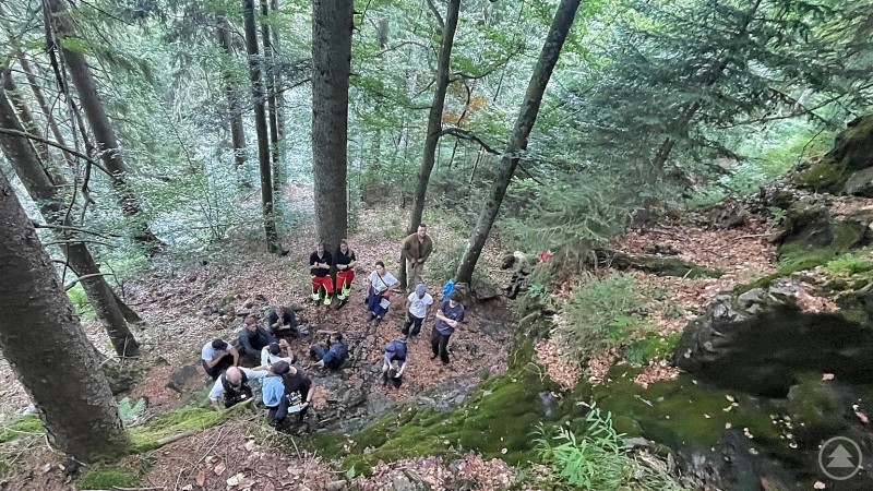 Filmteam und Schauspieler bei Dreharbeiten in den Wäldern nahe Bodenmais. Eine Gruppe von Menschen sitzt und steht in einem bewaldeten Hang mit Moos und Felsen, während sie eine Filmszene vorbereiten.