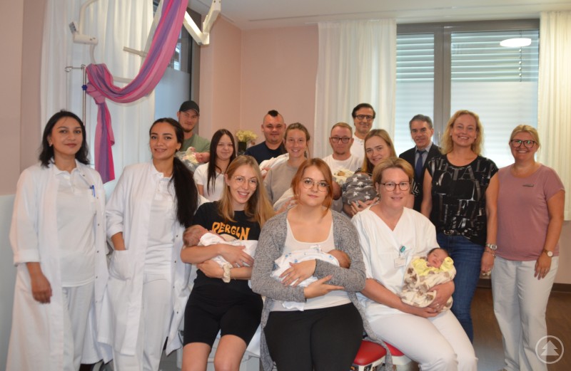 Chefarzt Sokol Rexhepi, Belegarzt Dr. Bernhard Rabenbauer, die Hebammen Kathrin Kirschner und Sylvia Stöckl sowie die Assistenzärztinnen freuen sich gemeinsam mit den jungen Familien über sieben Geburten in 24 Stunden am Krankenhaus Freyung. Eine große Gruppe von Menschen steht in einem hellen Raum. Vorne sitzen mehrere Mütter mit ihren Neugeborenen im Arm, dahinter stehen Väter, Ärztinnen, Hebammen und Krankenhausmitarbeitende. Alle lächeln in die Kamera.