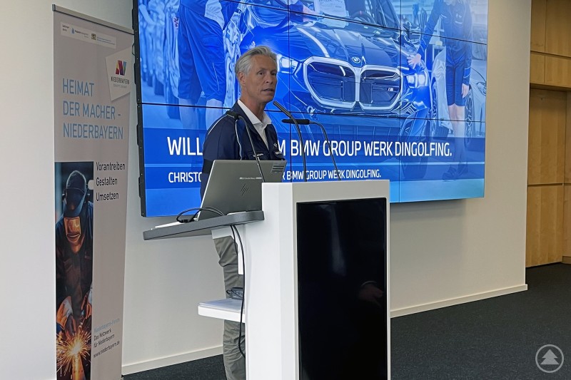 Christoph Schröder, Werkleiter des BMW Group Werks Dingolfing. Ein Mann steht an einem Rednerpult mit Mikrofon, hinter ihm eine große Leinwand mit der Aufschrift „BMW Group Werk Dingolfing“ und einem Bild eines BMW-Fahrzeugs.