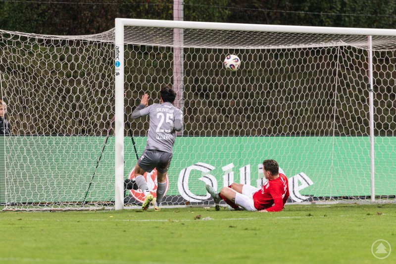 Ein Spieler im grauen Trikot trifft per Direktabnahme ins Tor, während ein Gegenspieler in Rot vergeblich versucht, den Ball auf der Linie zu klären.