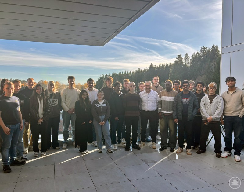 Eine Gruppe von Studierenden steht auf einer Terrasse mit modernem Gebäude und Blick auf den Bayerischen Wald im Hintergrund. Die Sonne scheint, und alle lächeln in die Kamera.