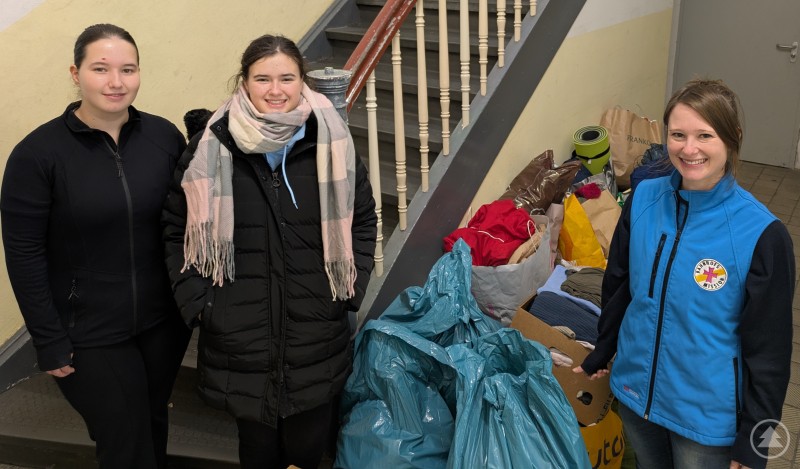 v.l.: Laura Witt (Schülerin), Luzie Wagner (Schülerin), rechts Julia Wawra (stellvertretende Leitung der Bahnhofsmission) Drei junge Frauen stehen neben mehreren großen Taschen und Kartons mit gespendeter Kleidung im Treppenhaus. Zwei tragen schwarze Winterjacken, eine (rechts) trägt eine blaue Weste mit Logo der Bahnhofsmission. Im Hintergrund sieht man eine Treppe.