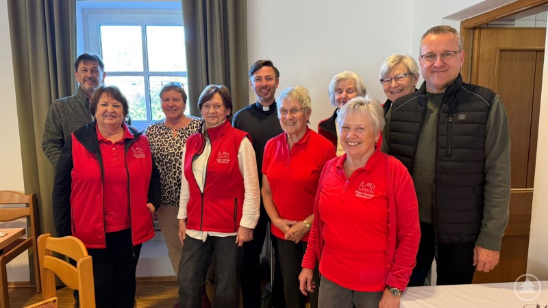 Das Organisationsteam des Frühstückstreffs für Alleinstehende in Vilshofen mit ehrenamtlichen Helferinnen, Verantwortlichen der Ortscaritas und Pfarrer Peter Kieweg. Eine Gruppe von Frauen und Männern steht lächelnd in einem Raum vor einem Fenster. Mehrere Frauen tragen rote Westen mit Caritas-Logo. In der Mitte steht ein Geistlicher, daneben Ehrenamtliche und Verantwortliche der Ortscaritas.