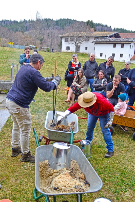 Hanne Juhre und Anita Springer in Aktion Das Bild zeigt eine lebendige Vorführung im Freien – vermutlich im Rahmen des Eiermarkts 2025 im Freilichtmuseum. Zwei Personen in Handschuhen und Schutzkleidung arbeiten konzentriert an einer traditionellen Handwerksaktion, vermutlich Metallguss oder Schmieden mit Sandformen, wie es die Schubkarren mit Sand und Blechteilen vermuten lassen. Interessierte Besucherinnen und Besucher – darunter Erwachsene und ein Kind im Bollerwagen – schauen aufmerksam zu. Im Hintergrund sind historische Gebäude und die bewaldete Hügellandschaft zu sehen. Die Szene vermittelt authentisches Handwerk, Wissensvermittlung und gemeinsames Erleben in ländlich-idyllischem Ambiente.