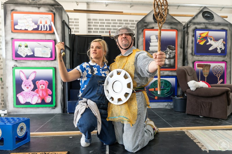 Zwei Schauspieler in fantasievollen Kostümen posieren auf einer Bühne – mit Alltagsgegenständen als Requisiten. Im Hintergrund eine bunte Kulisse mit kindlichen Motiven.