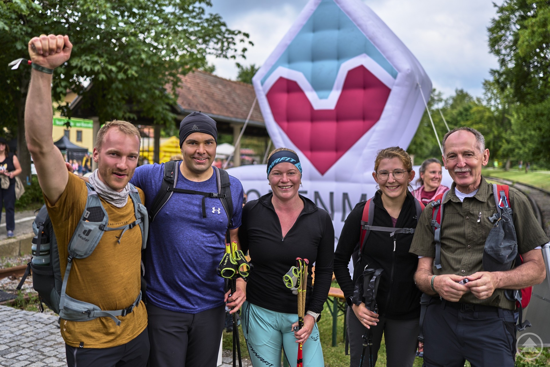 Fünf Wanderer posieren lächelnd und erschöpft vor einem Veranstaltungsbanner nach erfolgreicher Teilnahme an einer Wandertour.
