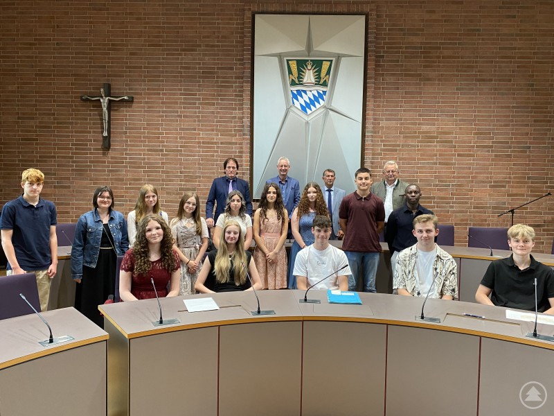 Die geehrten Schülerinnen und Schüler gemeinsam mit Landrat Josef Laumer, Oberbürgermeister Markus Pannermayr und den Schulamtsdirektoren Heribert Ketterl und Stephan Grotz. Eine Gruppe junger Menschen sitzt und steht im großen Sitzungssaal eines Landratsamtes. Hinter ihnen sind ein bayerisches Wappen, ein Kreuz und eine Wand aus roten Ziegelsteinen zu sehen. Erwachsene Vertreter aus Politik und Schule stehen in der zweiten Reihe.
