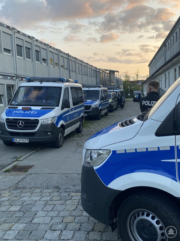 Eine Reihe von Polizeibussen steht in einem schmalen Innenhof zwischen zwei langgestreckten Gebäuden bei Sonnenaufgang.