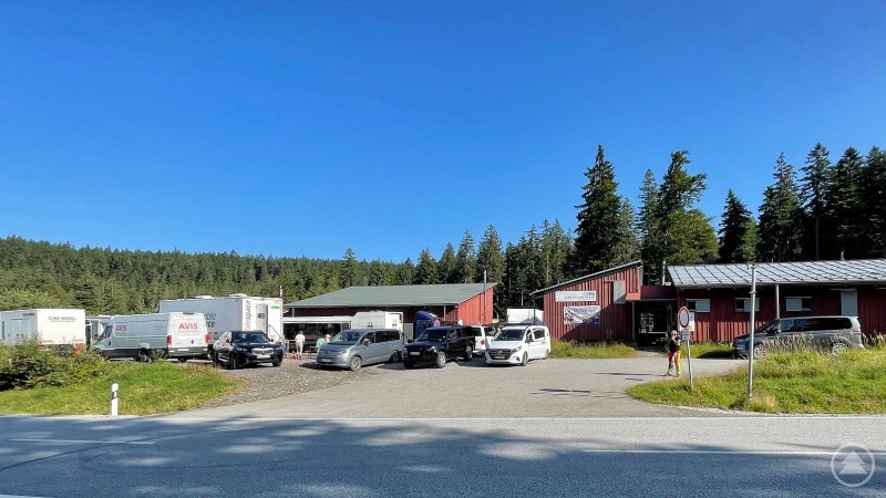 Mehrere Transportfahrzeuge und LKWs stehen vor roten Gebäuden, die als Basislager dienen, umgeben von Wald und blauem Himmel.