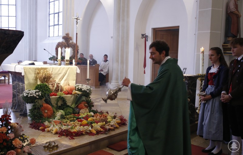 Pfarrer Simon Steinbauer segnete die Gaben der Gläubigen und erinnerte daran, achtsam mit der Schöpfung umzugehen. Pfarrer Simon Steinbauer segnet das Erntedank-Arrangement mit Weihrauch, während Gläubige in Tracht am Altar stehen.