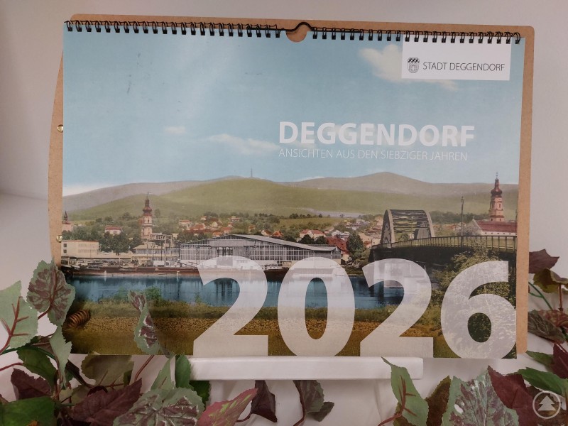 Der neue Fotokalender 2026 der Stadt Deggendorf zeigt historische Aufnahmen aus den 1970er-Jahren. Ein Wandkalender der Stadt Deggendorf mit dem Titel „Deggendorf – Nostalgie in Ostbayerns Hauptstadt 2026“ steht auf einem weißen Ständer, umrahmt von grünen und roten Blättern. Auf dem Titelblatt ist ein historischer Blick auf Deggendorf mit Brücke und Stadtpanorama zu sehen.