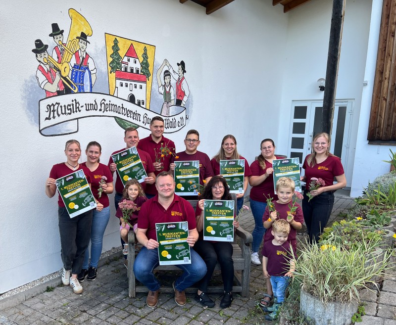 Das Bild zeigt eine fröhliche Gruppe von Mitgliedern des Musik- und Heimatvereins Haus i. Wald e.V., die gemeinsam vor dem Vereinsheim posieren. Alle tragen bordeauxrote Vereins-T-Shirts und halten Plakate in der Hand, die auf das „1. Musikantentreffen“ hinweisen. Einige Mitglieder haben zudem Blumensträuße dabei, und auch Kinder sind mit auf dem Foto. Im Hintergrund ist das Vereinslogo mit Musikantenmalerei an der Wand gut sichtbar.