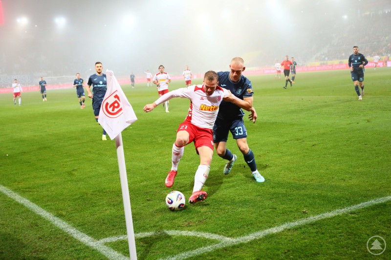 Ein Spieler des SSV Jahn Regensburg kämpft an der Eckfahne mit einem Münchner Verteidiger um den Ball, während mehrere Mitspieler und der Schiedsrichter im Hintergrund stehen.