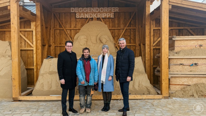 Deutschlands größte Sandkrippe entsteht im Jahr 2025 bereits zum dritten Mal und wird durch einen zweiten Anbau noch größer: v.l. Oberbürgermeister Dr. Christian Moser, Sandkünstler Jiří Kašpar, Lisa Peter vom Stadtmarketing und Stadtmarketing Vorstand Andreas Höhn Vier Personen stehen vor einer teilweise fertig geformten großen Sandkrippe in einer Holzbaukonstruktion; im Hintergrund sind Sandblöcke, Werkzeuge und die Beschriftung „Deggendorfer Sandkrippe“ zu sehen.