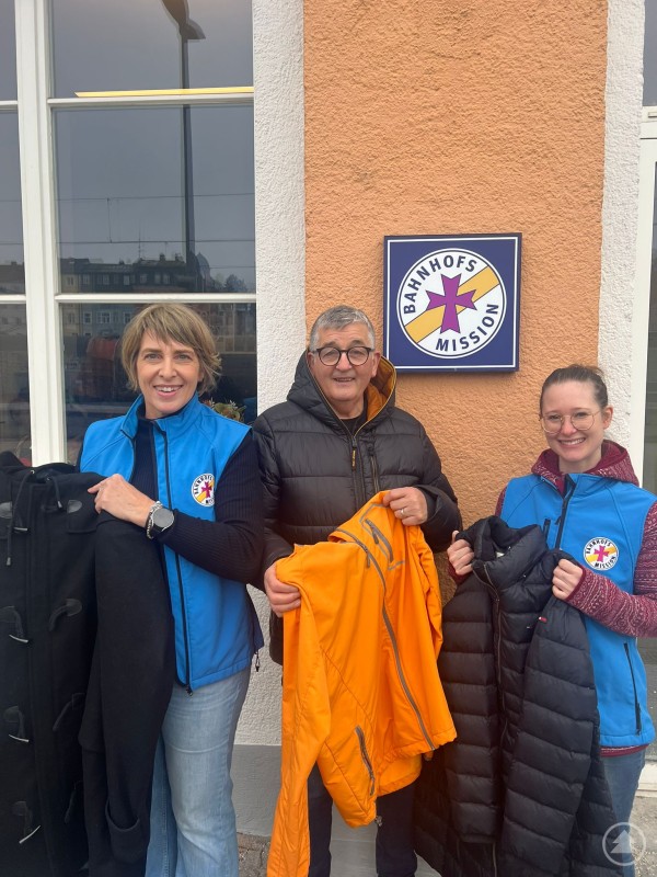 v.l.: Angelika Leitl-Weber (Einrichtungsleiterin der Bahnhofsmission), Hans Weber (Inhaber BIGDANDY), Julia Wawra (stellv. Leiterin der Bahnhofsmission) Drei Personen stehen vor dem Eingang der Bahnhofsmission Passau und halten warme Winterjacken in den Händen. Zwei Frauen tragen blaue Westen mit dem Logo der Bahnhofsmission, der Mann in der Mitte hält eine auffällige orange Jacke. Im Hintergrund ist das Schild der Bahnhofsmission an der Hauswand sichtbar.