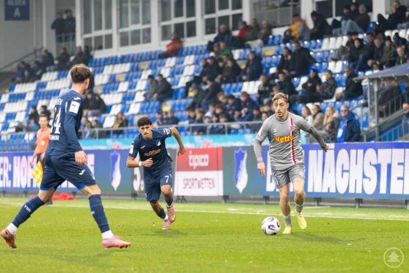 Die Jahnelf setzt zum Angriff an, Hoffenheim verteidigt aufmerksam. Ein Spieler des SSV Jahn dribbelt mit dem Ball, während zwei Hoffenheimer Spieler anlaufen.