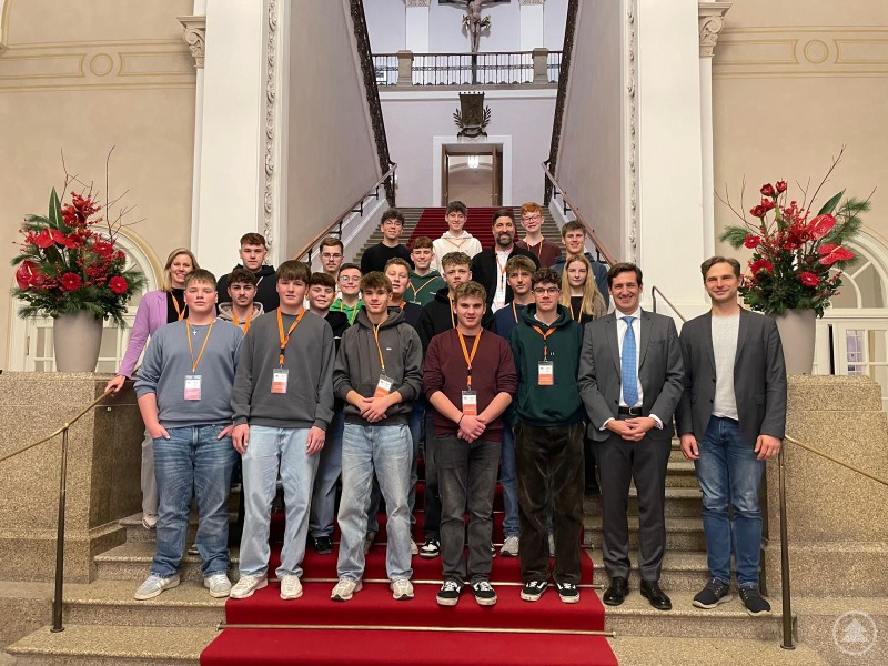 Die Abgeordneten des Wahlkreises Toni Schuberl, Dr. Stefan Ebner und Martin Behringer (nicht im Bild) nahmen sich bei einem Besuch des Maximilianeums ausführlich Zeit für einen direkten Austausch mit den Grafenauer Realschülerinnen und –schülern und deren Lehrkräften Sabine Pinter und Ferdinand Klingelhöfer. Eine Schulklasse mit Lehrkräften steht im Inneren des Maximilianeums auf der großen Treppe. Die Jugendlichen und Erwachsenen sind gruppiert auf rotem Teppich, im Hintergrund sind Säulen und florale Dekorationen zu sehen.