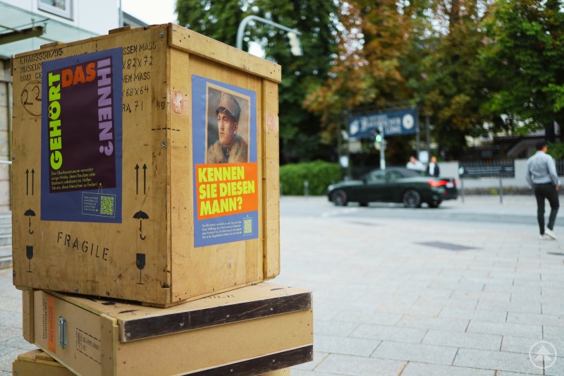 Kunsttransportkisten mit Plakaten ungeklärter Kunstwerke auf dem Ludwigsplatz in Passau machten die Provenienzforschung des Oberhausmuseums für die Öffentlichkeit sichtbar. Holzkisten für Kunsttransporte stehen im öffentlichen Raum am Ludwigsplatz in Passau. Auf den Kisten sind Plakate mit historischen Porträts und der Frage Kennen Sie diesen Mann? angebracht. Im Hintergrund ist eine Straße mit Passanten und Fahrzeugen zu sehen.