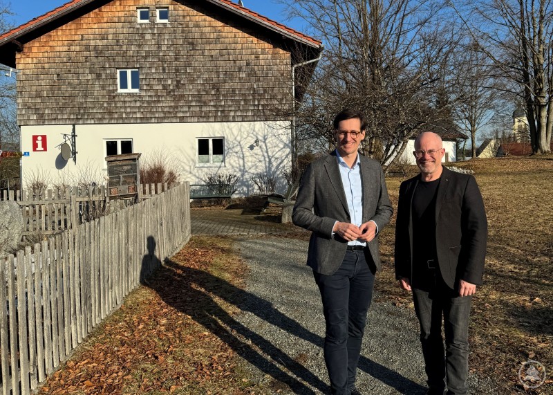 Trafen sich zum Austausch über die Ferienregion Nationalpark Bayerischer Wald an der Geschäftsstelle in Spiegelau: Dr. Stefan Ebner MdL links und Robert Kürzinger. Zwei Männer stehen vor einem Gebäude im Freien und unterhalten sich. Im Hintergrund ist eine ländliche Umgebung mit Zaun, Wiese und Bäumen zu sehen.
