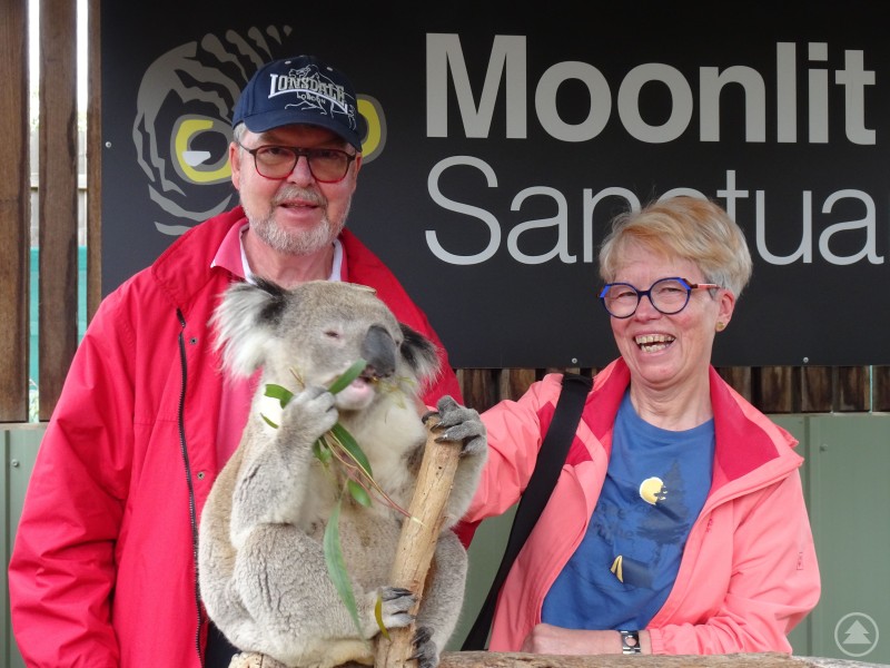Roland Walter und Ingeborg Endl-Walter präsentieren ihre Reiseeindrücke aus Australien. Zwei Personen stehen vor einem Schild und halten gemeinsam einen Koala, der an einem Ast frisst.