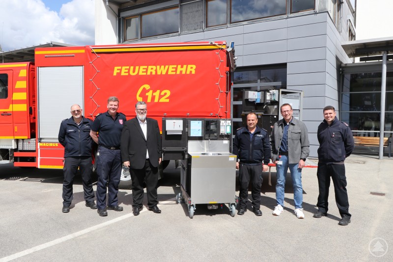 Bürgermeister Andreas Rother (3. v. l) zusammen mit (v. l) dem Wachleiter der Hauptamtlichen Tagwache, Alexander Kornexl, Stadtbrandrat Andreas Dittlmann, dem Kommandanten der Feuerwehr Heining, Markus Freundorfer, dem stellv. Kommandanten der Feuerwehr Heining, Severin Putz sowie dem Dienststellenleiter des Amtes für öffentliche Ordnung, Erik Linseisen, bei der Übergabe der beiden neuen Hygiene Rollcontainer für die Feuerwehren Hauptwache und Heining. Sechs Personen stehen vor einem Feuerwehrfahrzeug mit der Aufschrift Feuerwehr 112. Neben ihnen ist ein mobiler Hygiene Rollcontainer zu sehen, der offiziell übergeben wird.