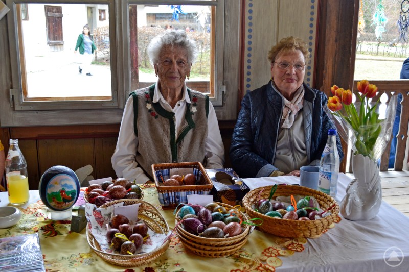 Antschi Aumüller älteste Teilnehmerin Das Bild zeigt zwei ältere Damen an einem liebevoll dekorierten Stand beim Eiermarkt 2025. Sie sitzen in einem traditionellen Raum mit bemalten Wänden und blicken freundlich in die Kamera. Vor ihnen auf dem Tisch liegen kunstvoll verzierte Ostereier in geflochtenen Körben – teils gefärbt, teils mit feinen Mustern versehen. Daneben stehen Getränke, ein Strauß frischer Tulpen sowie ein großer, bemalter Zier-Ei-Ballon. Die Szene vermittelt Herzlichkeit, Handwerkskunst und die Bewahrung regionaler Traditionen – ein authentischer Moment bäuerlicher Osterkultur in gemütlicher Atmosphäre.