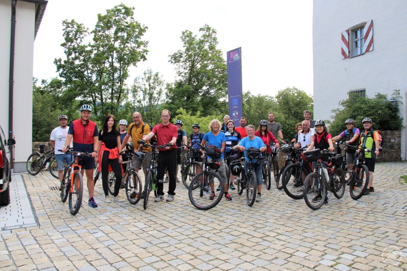 Landrat Sebastian Gruber (2. v. links) hofft auf rege Beteiligung bei der Feierabend-Radltour im Rahmen der STADTRADELN-Kampagne. Eine Gruppe von Radfahrerinnen und Radfahrern steht mit ihren Fahrrädern auf einem gepflasterten Platz vor dem Schloss Wolfstein. Die Teilnehmenden lächeln in die Kamera, viele tragen Helme und Sportkleidung.