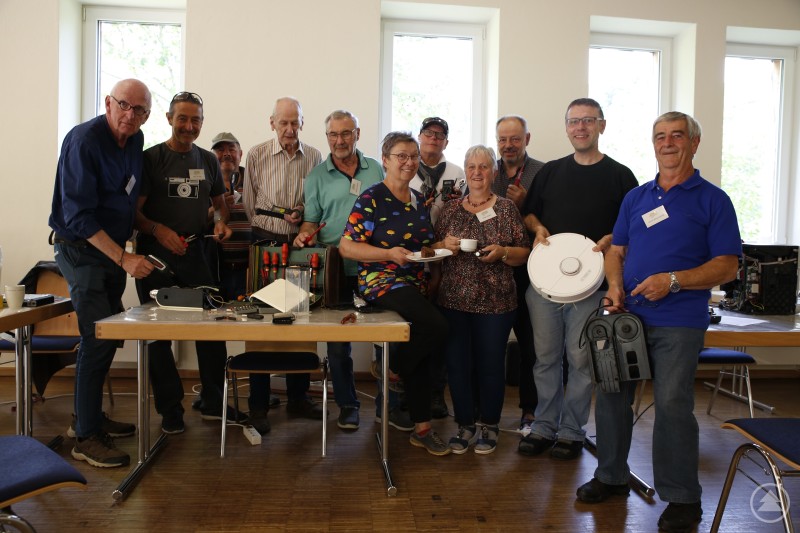 Das Team für Kabelbrüche und lockere Schrauben hat jetzt 12 Reparatur-Café-Termine hinter sich gebracht und ist weiter mit jeder Menge Herzblut und Lötzinn am Elektro-OP-Tisch zu Gange: Ron Kulik (v.l.), Peter Schöpp, Georg Pichler, Heinz-Dieter Schubardt, Manfred Duschl, Heidi Leutner, Kristian Seinsch, Ulrike Hilpert, Rudolf Leutner, Martin Schweighofer, Armin Braumandl. Foto: Markt Schönberg Eine Gruppe von etwa einem Dutzend Menschen steht in einem Raum mit Werkzeugen, defekten Geräten und Kaffeetassen, einige halten Reparaturgegenstände in der Hand.