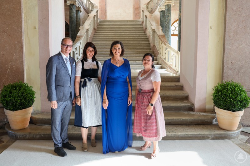 Während Ilse Aigner elegant in blauem Kleid für mehr Frauen in Politik und Ehrenamt warb, fühlten sich die Damen in Tracht Nadine Aulinger (r.) sowie Nicole Polleichtner in ihrem Engagement bereits gerne bestätigt. MdL Martin Behringer durfte „assistieren“. Vier Personen posieren im Eingangsbereich eines barocken Schlosses, zwei Frauen tragen Dirndl, eine Frau ein elegantes blaues Kleid, ein Mann einen grauen Anzug.