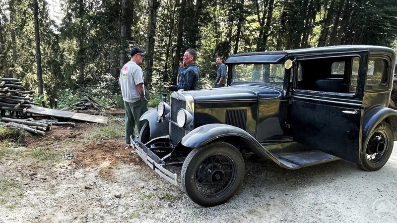 Oldtimer-Szene beim Filmdreh von „THE WEIGHT“ in den Wäldern bei Bodenmais. Ein Oldtimer steht auf einem Waldweg, daneben sprechen drei Männer, während Holzstapel im Hintergrund zu sehen sind.