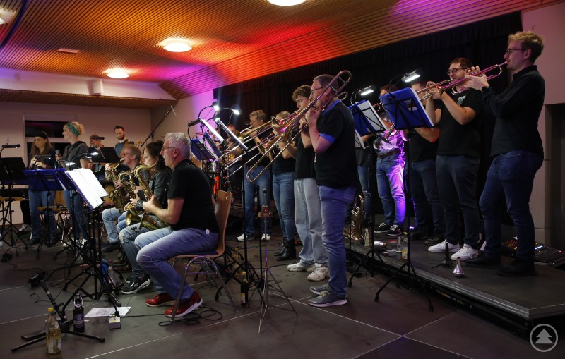 Mit einem Jahr Verspätung, aber kräftig bei Puste, blies Makapeo den Konzertbesuchern im KUK den Funk, R&B, Pop und Metal um die Ohren. Die Big Band „Makapeo“ der Marktkapelle Obernzell steht auf einer Bühne im KUK Schönberg. Musikerinnen und Musiker spielen mit Trompeten, Posaunen, Saxophonen und Rhythmusinstrumenten unter farbigem Bühnenlicht.