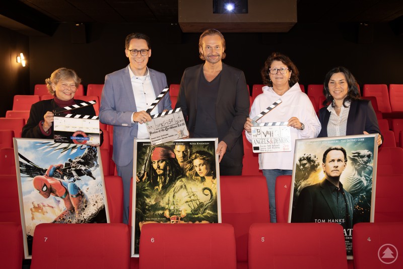 Hela Schandelmaier, Oberbürgermeister Dr. Christian Moser, Kameramann Nino Neuböck, Kinobetreiberin Eva Schattenfroh und Petra Schwankl freuen sich auf den Kinoabend des Familienzentrums und der Stadt Deggendorf im Lichtspielhaus. Fünf Personen stehen in einem Kinosaal mit roten Sitzen. Sie halten Filmklappen und große Filmplakate zu bekannten Hollywood-Produktionen wie „Spider-Man“, „Fluch der Karibik“ und „Illuminati“. Im Hintergrund ist ein laufender Projektor zu sehen, der eine Kinoatmosphäre schafft.