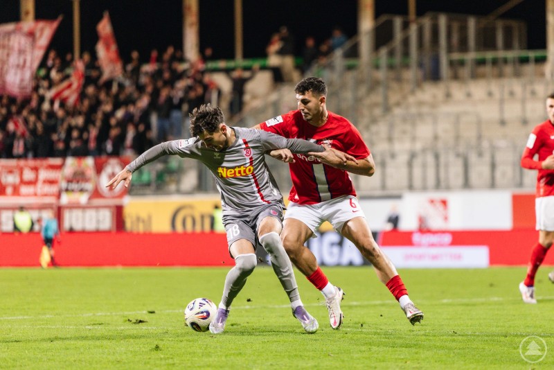 Kampf um jeden Ball – intensives Duell zwischen Jahn Regensburg und dem TSV Havelse. Ein Spieler im grauen Trikot behauptet den Ball im Zweikampf gegen einen Gegenspieler in Rot, während im Hintergrund Fans mit Fahnen jubeln.