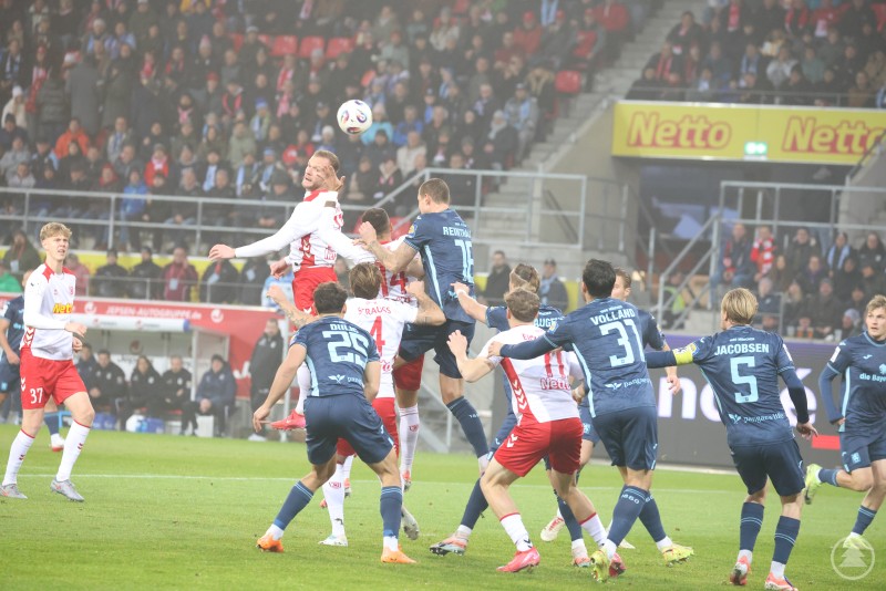 Mehrere Spieler des SSV Jahn Regensburg und des TSV 1860 München springen im Strafraum hoch, um den Ball per Kopf zu erreichen. Im Hintergrund verfolgen zahlreiche Zuschauer die Szene gespannt.