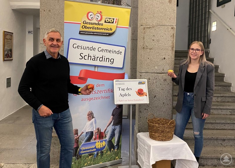 Bürgermeister Günter Streicher und die zuständige Stadtgemeinde-Mitarbeiterin Jana Jungbauer. Zwei Personen stehen in einem Eingangsbereich neben einem Informationsbanner der „Gesunden Gemeinde Schärding“. Beide halten einen Apfel in der Hand, daneben steht ein Korb auf einem kleinen Tisch.