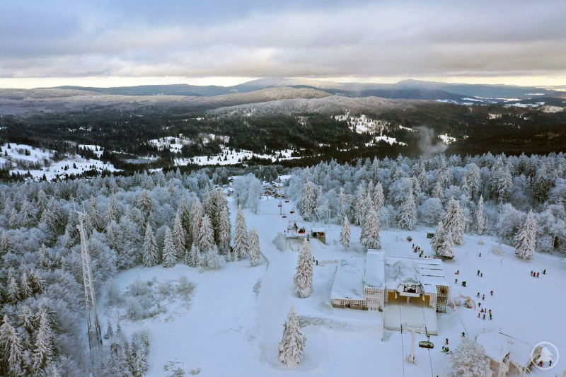 Blick über den Almberg im Skizentrum Mitterdorf: Die neuen Liftanlagen prägen das modernisierte Erscheinungsbild des Skigebiets und stärken den Standort nachhaltig. Luftaufnahme des verschneiten Almbergs mit den neuen Sesselbahnen, Talstation und umliegenden Wäldern im Winter.