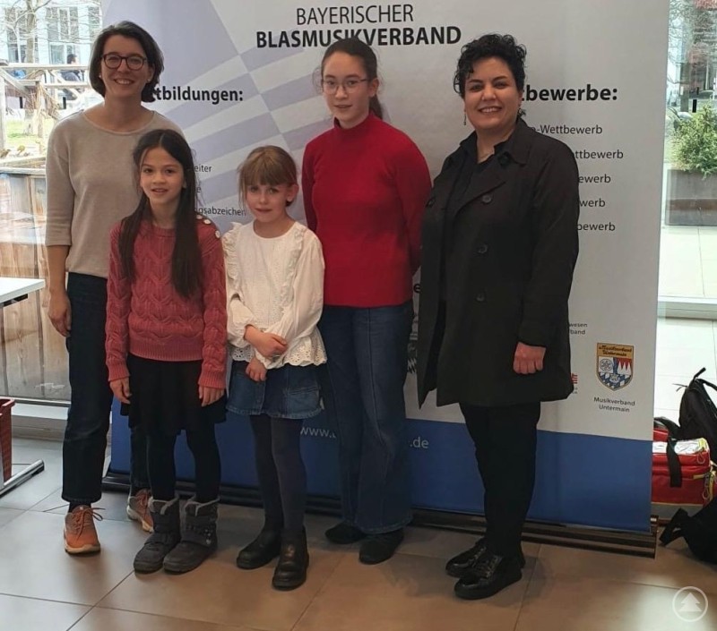 Über den souveränen Auftritt in Nürnberg freuten sich Lehrkräfte und Schülerinnen gleichermaßen (von links): Elisabeth Wagner-Thoma, Amelie Hartl, Antonia Burkert, Mária Król und Setareh Shafii Tabatabai. Vor dem Banner des Bayerischen Blasmusikverbands stehen fünf Personen in einer hellen Eingangshalle nebeneinander und lächeln in die Kamera. Zwei Erwachsene – vermutlich Lehrkräfte – flankieren drei junge Mädchen, die als Teilnehmerinnen des „Concertino“-Landesentscheids aufgetreten sind. Die Kinder tragen winterliche Kleidung, während der Hintergrund das Logo und Informationen zu Wettbewerben und Ausbildungen im Blasmusikverband zeigt. Die freundliche Atmosphäre deutet auf einen erfolgreichen und freudigen Anlass hin.